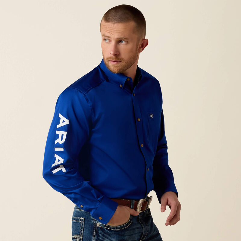 Chambelán look: Ariat Team Logo twill shirt in ultramarine blue with white lettering on the sleeve, worn with jeans and a western belt, for quinceañeras and weddings in Dallas